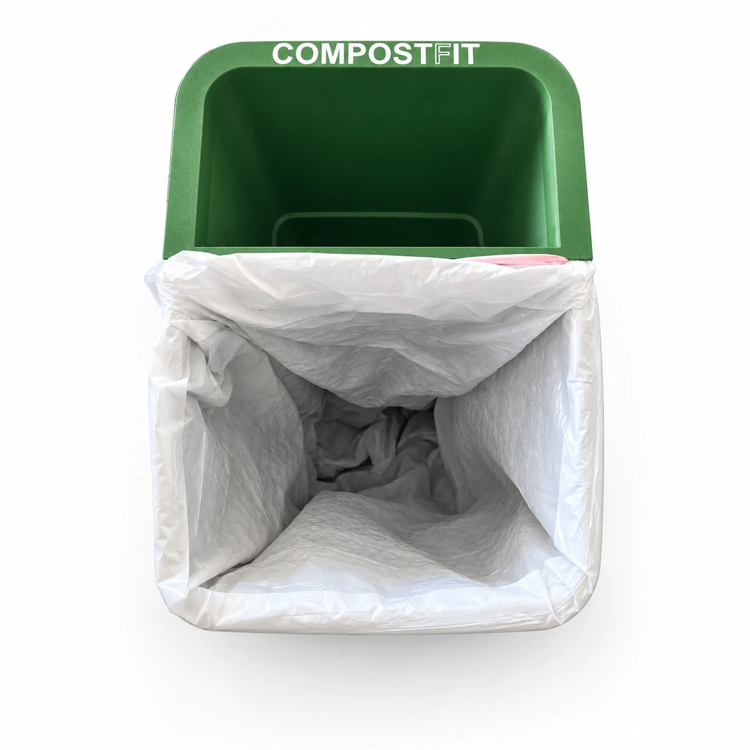 Top-down view of CompostFit seated in a pull-out trash can, the white trash bag swept around it and gripped under the green COMPOSTFIT rim